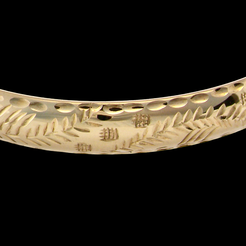 A close-up of a polished yellow gold bangle bracelet featuring engraved leaf and branch motifs, set against a solid black background.