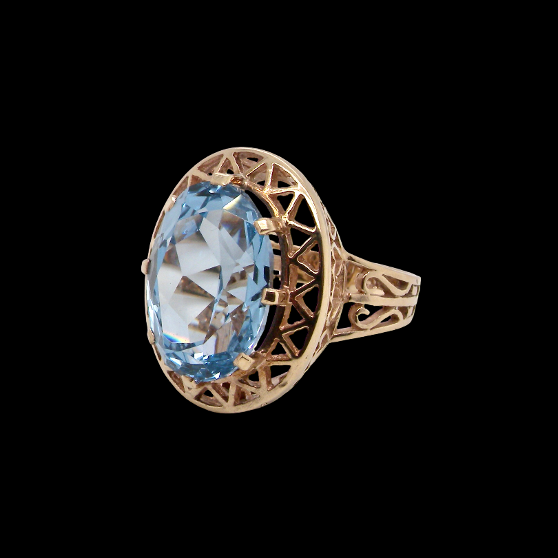 An antique-style gold ring featuring a large oval-cut light blue gemstone, set in an openwork filigree band, shown against a solid black background.
