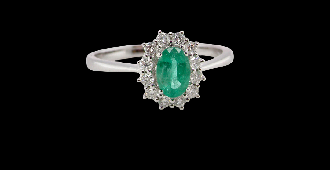 A classic cluster ring set with an oval cut emerald centre stone, measures 6x4mm, grass green colour.
Complimented by an outer halo of twelve brilliant cut white diamonds,