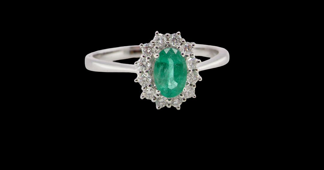A classic cluster ring set with an oval cut emerald centre stone, measures 6x4mm, grass green colour.
Complimented by an outer halo of twelve brilliant cut white diamonds,
