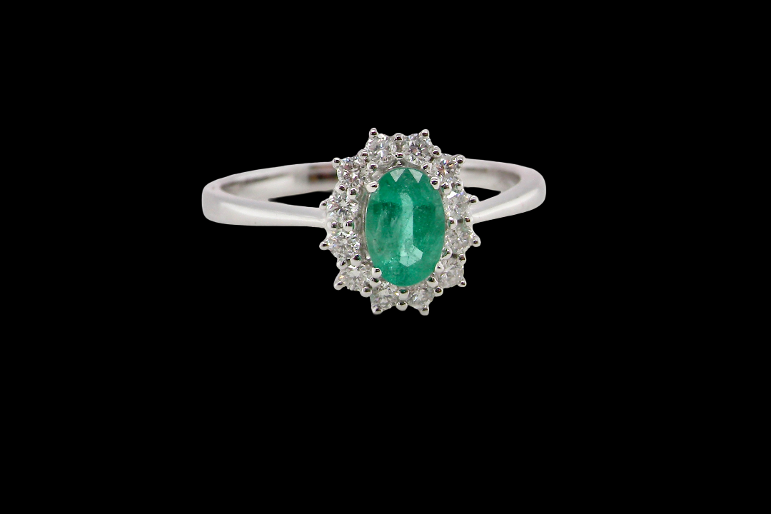 A classic cluster ring set with an oval cut emerald centre stone, measures 6x4mm, grass green colour.
Complimented by an outer halo of twelve brilliant cut white diamonds,