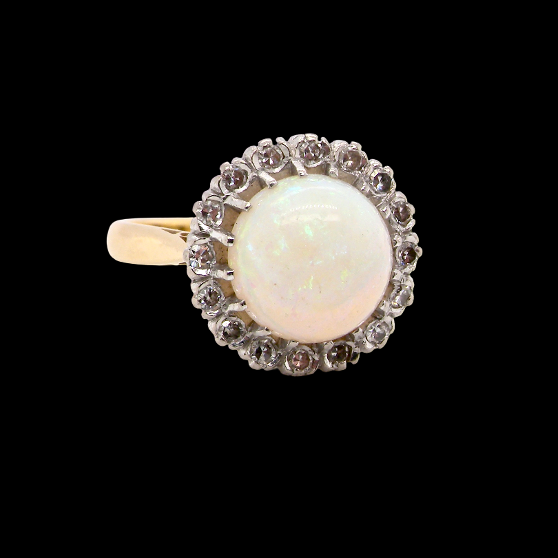 A vintage-style ring featuring a round white opal center stone surrounded by a halo of small clear stones, set on a yellow gold band, photographed against a solid black background.