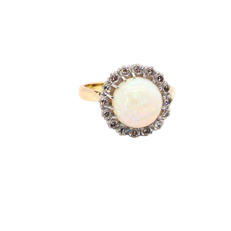 A vintage-style ring featuring a round white opal center stone surrounded by a halo of small dark stones, set on a yellow gold band, isolated on a white background.