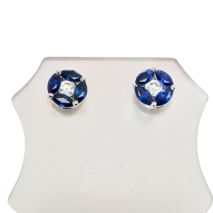 A pair of round stud earrings with deep blue gemstones arranged in a flower shape around a clear central stone, set in silver-toned metal, displayed on a white stand against a white background.