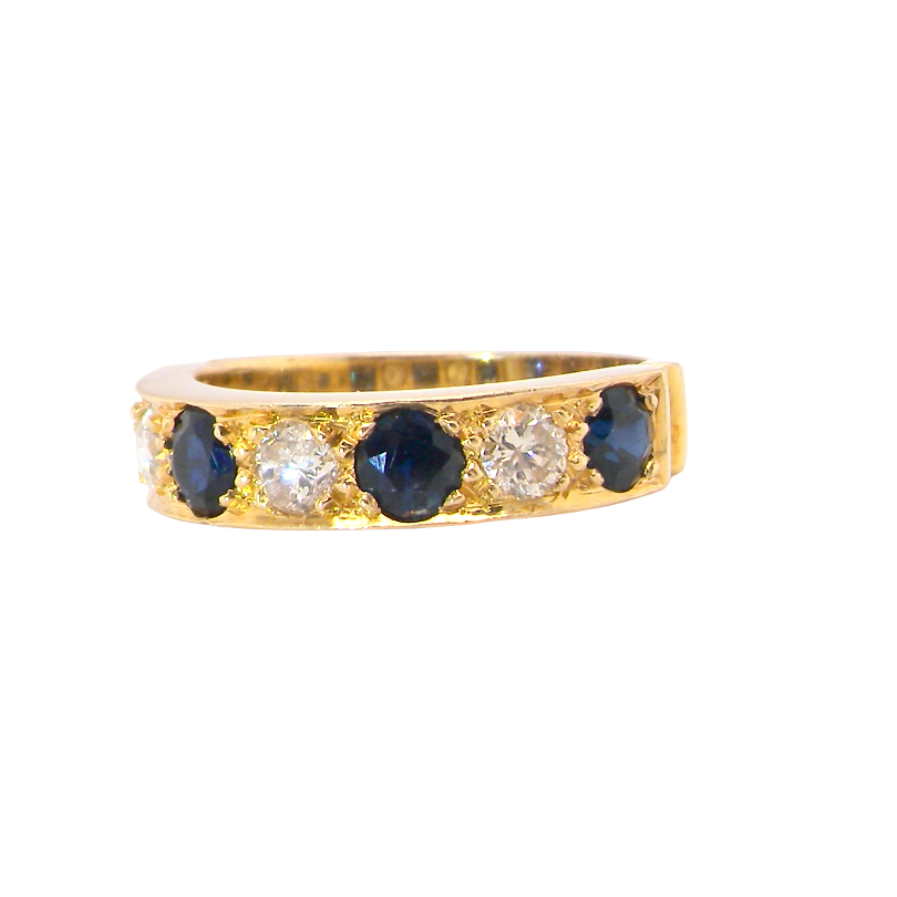 A yellow gold eternity-style ring featuring alternating round dark blue sapphires and clear round diamonds, shown against a plain white background.