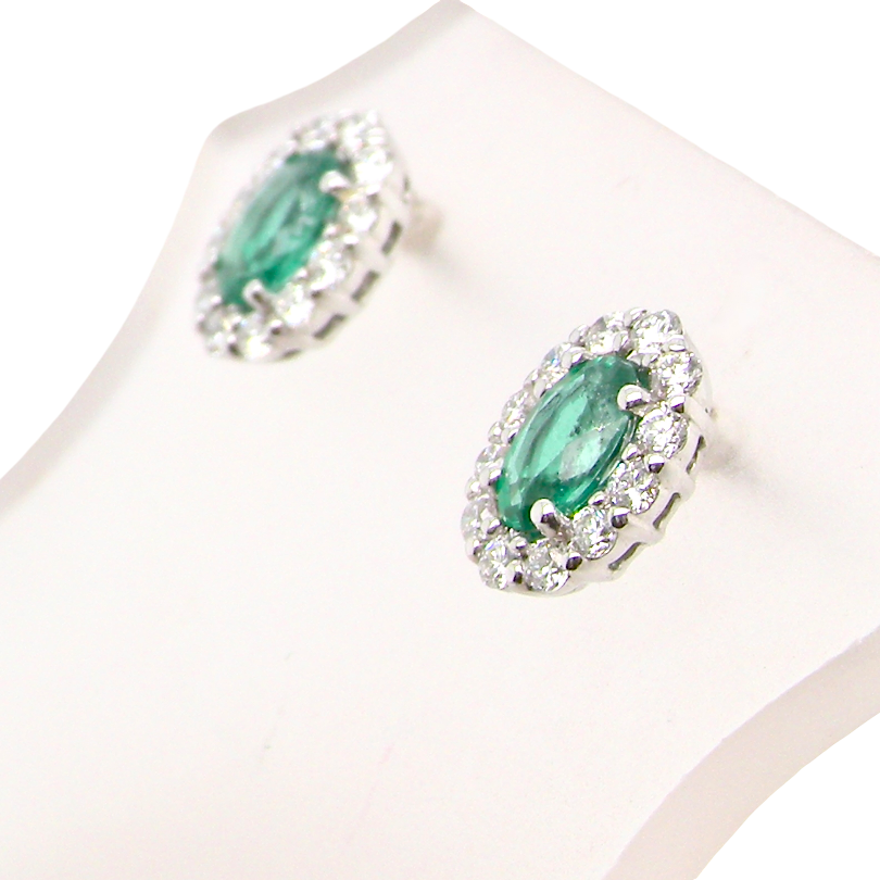 A pair of oval emerald-cut green gemstone stud earrings with silver-toned halo settings and small clear stones, displayed on a white jewelry stand against a white background.