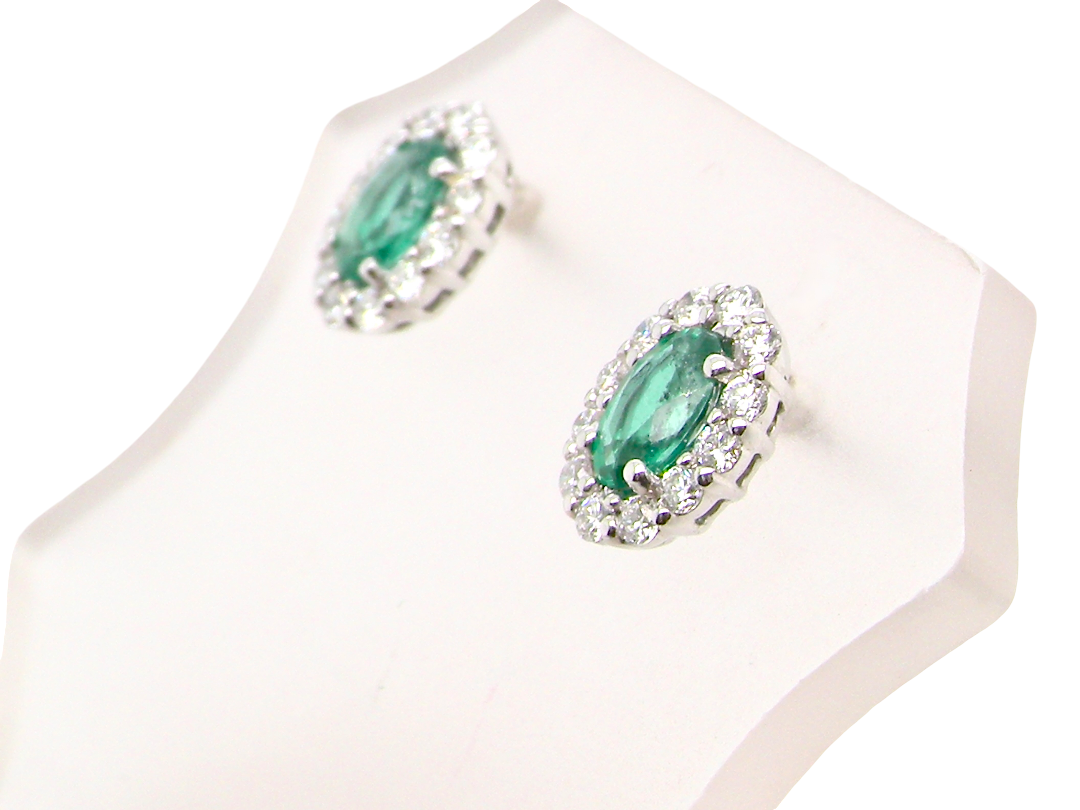 A pair of oval emerald-cut green gemstone stud earrings with silver-toned halo settings and small clear stones, displayed on a white jewelry stand against a white background.