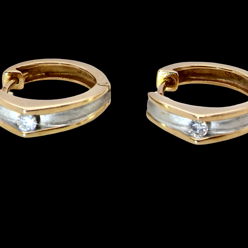 A pair of gold huggie hoop earrings with a silver inlay and a small clear gemstone on each earring, shown against a solid black background.