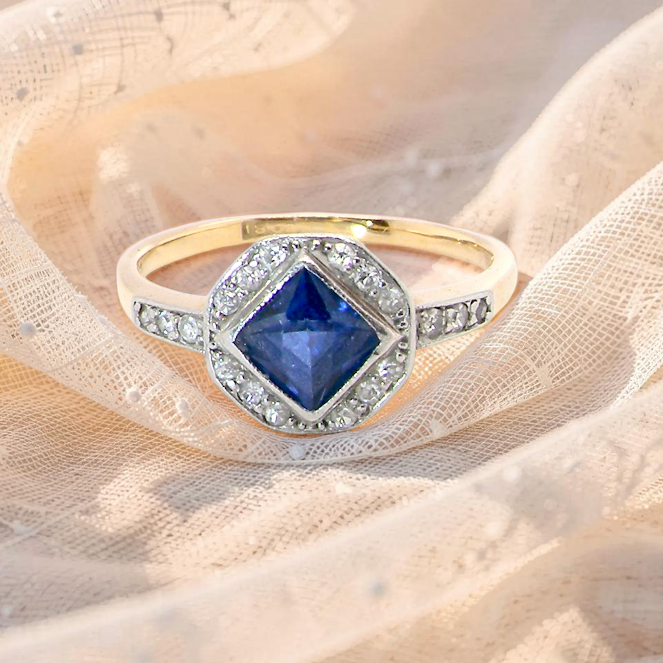 A very pretty Art Deco cluster ring. To the centre a mid to dark blue sapphire, square cut but set as a 'diamond shape'. Measures 5x5mm,