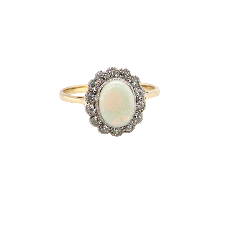 A vintage-style ring featuring an oval cabochon opal surrounded by a halo of small clear stones, set on a yellow gold band, isolated on a white background.