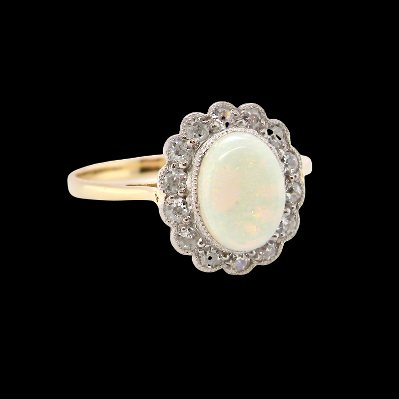 A yellow gold ring featuring an oval white opal center stone surrounded by a halo of small clear stones, set against a solid black background.