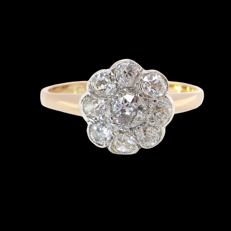 A vintage-style diamond cluster ring featuring a central round diamond surrounded by eight smaller diamonds, set in a yellow gold band against a solid black background.