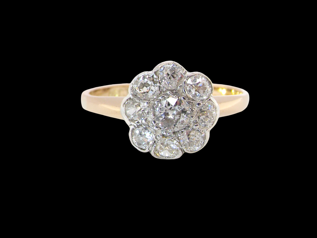 A vintage-style diamond cluster ring featuring a central round diamond surrounded by eight smaller diamonds, set in a yellow gold band against a solid black background.
