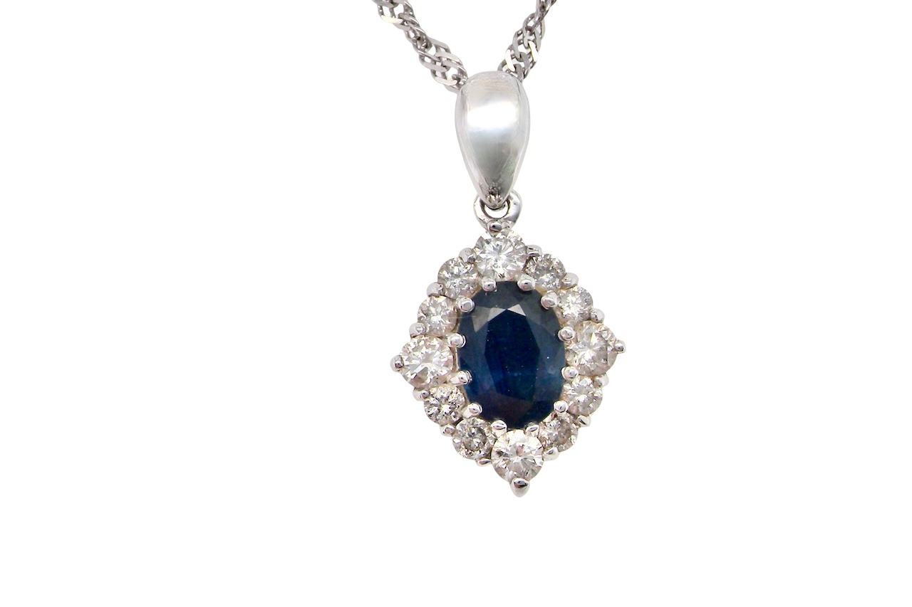 A white gold pendant. The centre stone an inky black blue sapphire 7x5mm, claw set. Complimented by an outer halo of brilliant cut white diamonds.