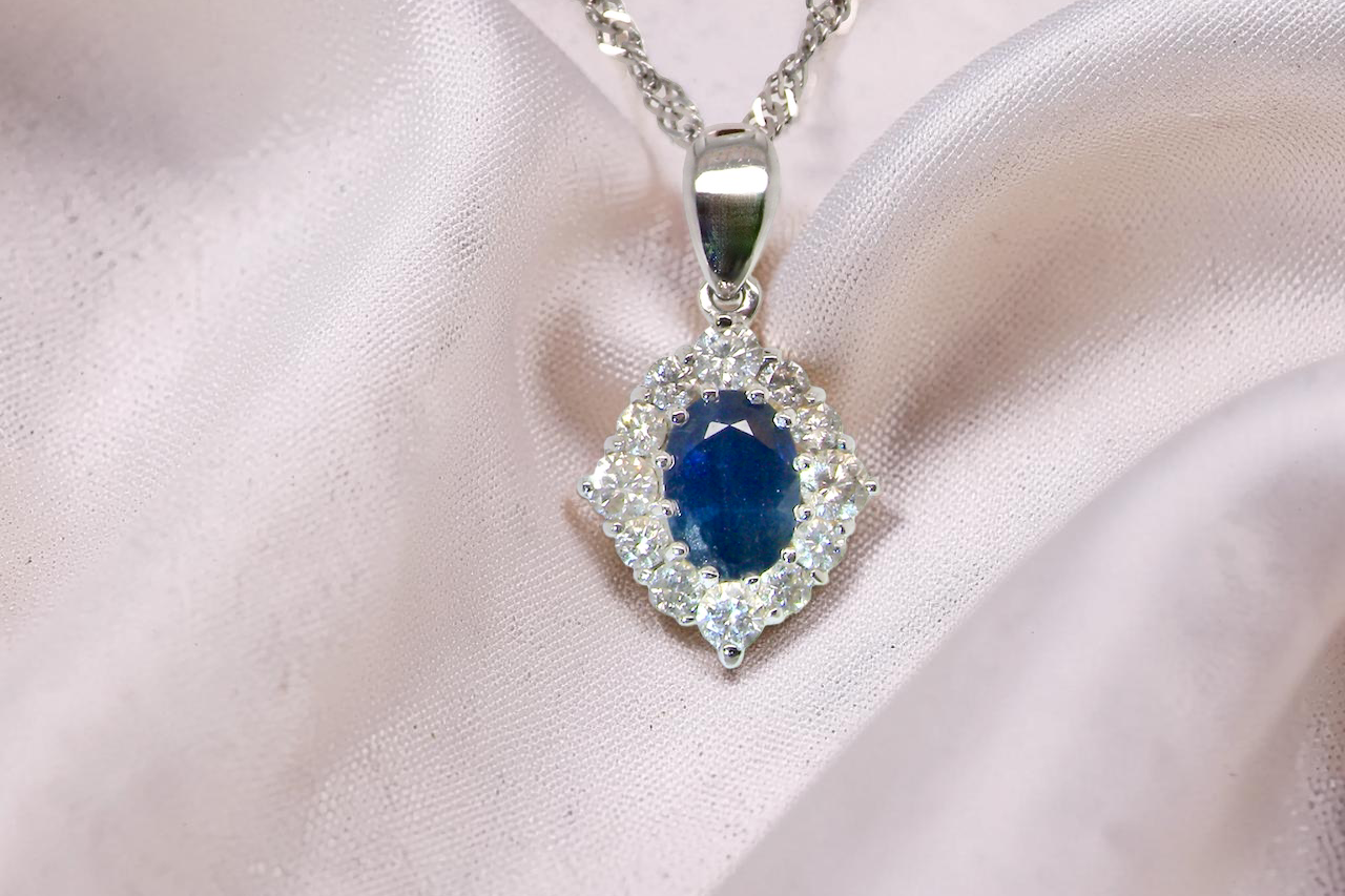 A white gold pendant. The centre stone an inky black blue sapphire 7x5mm, claw set. Complimented by an outer halo of twelve diamonds.