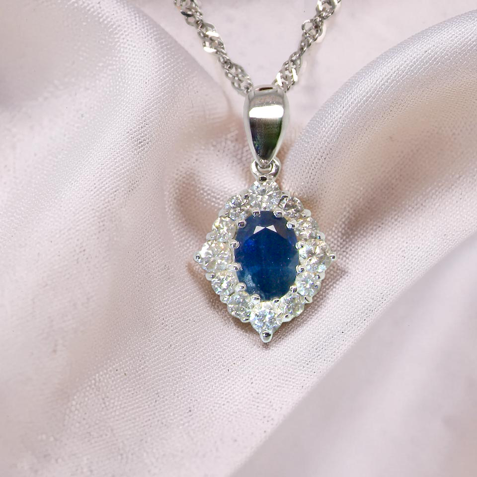A white gold pendant. The centre stone an inky black blue sapphire 7x5mm, claw set. Complimented by an outer halo of twelve diamonds.