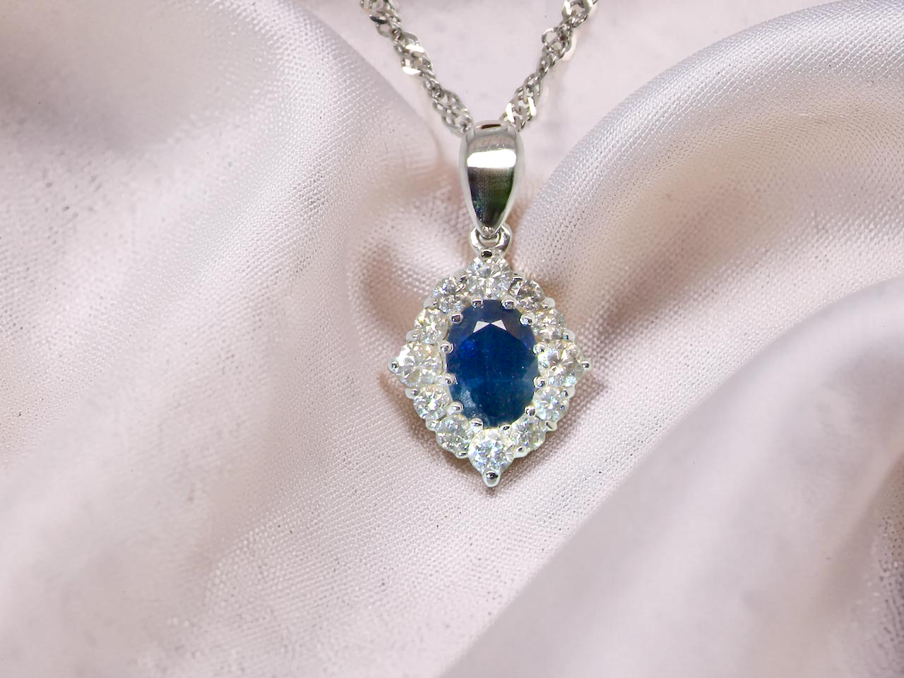 A white gold pendant. The centre stone an inky black blue sapphire 7x5mm, claw set. Complimented by an outer halo of twelve diamonds.