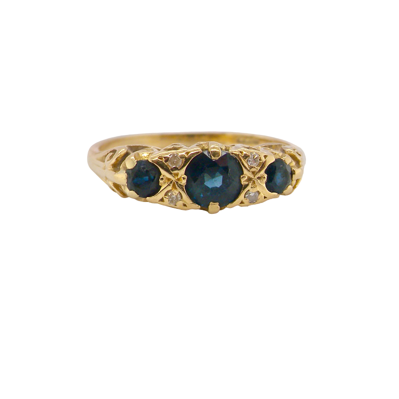 A vintage-style yellow gold ring featuring a central heart-shaped dark blue gemstone flanked by two smaller round blue gemstones, accented with small clear crystals, shown on a plain white background.