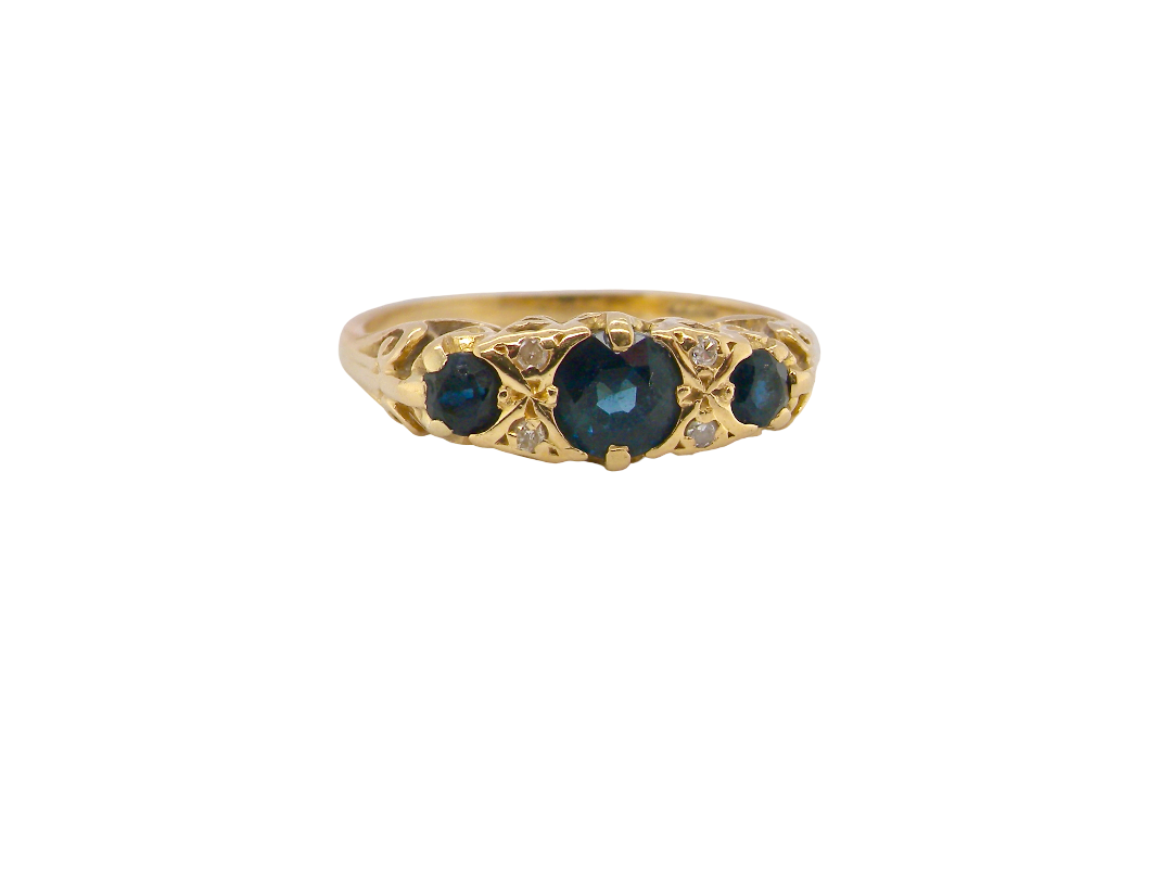 A vintage-style yellow gold ring featuring a central heart-shaped dark blue gemstone flanked by two smaller round blue gemstones, accented with small clear crystals, shown on a plain white background.
