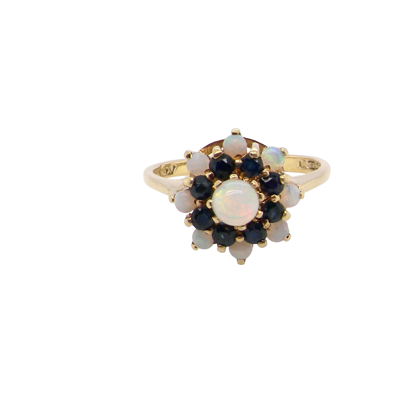 A gold ring featuring a flower-shaped cluster with a central round iridescent opal, surrounded by dark blue sapphires and smaller white opals, set against a plain white background.
