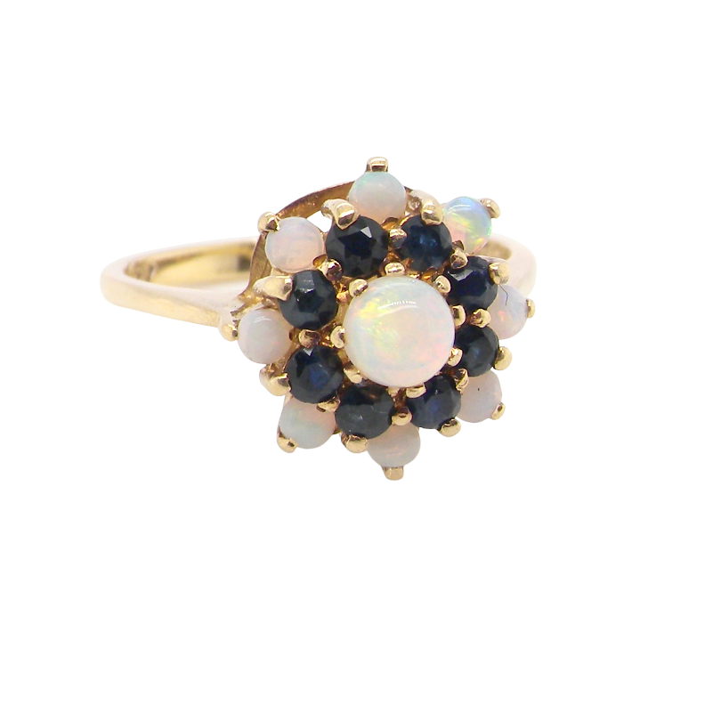 A gold ring featuring a flower-shaped cluster with a central oval white opal surrounded by dark blue sapphires and smaller round white opals, set against a plain white background.