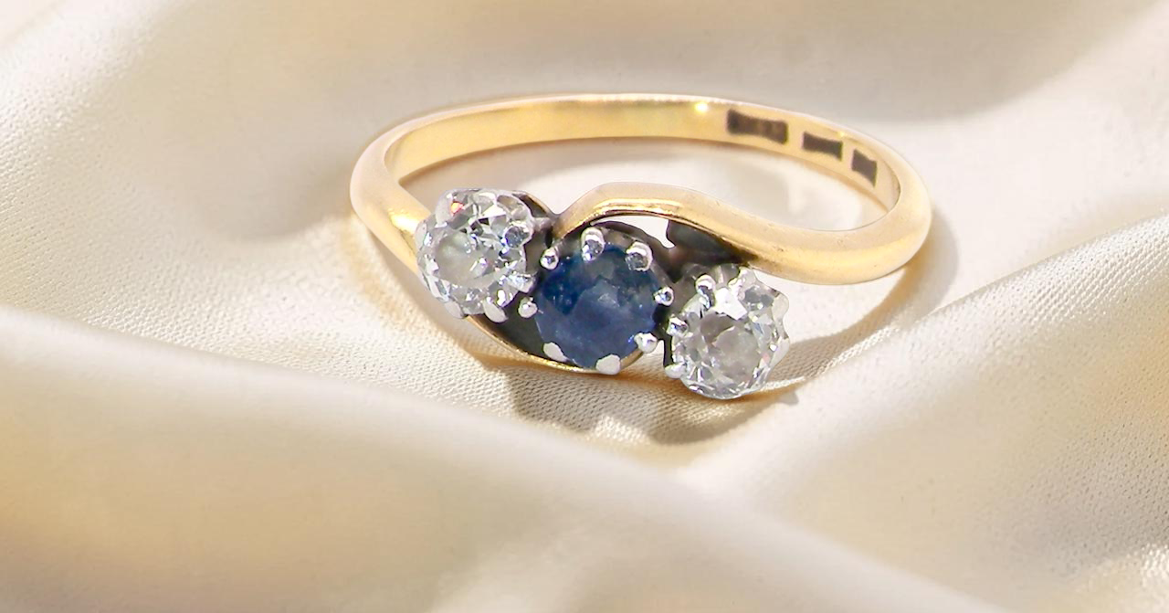 An 18 carat gold three stone cross over ring.
Centre stone a round sapphire 4mm diameter. Each side a brilliant cut white diamond 4mm diameter (0.25 carats estimated),