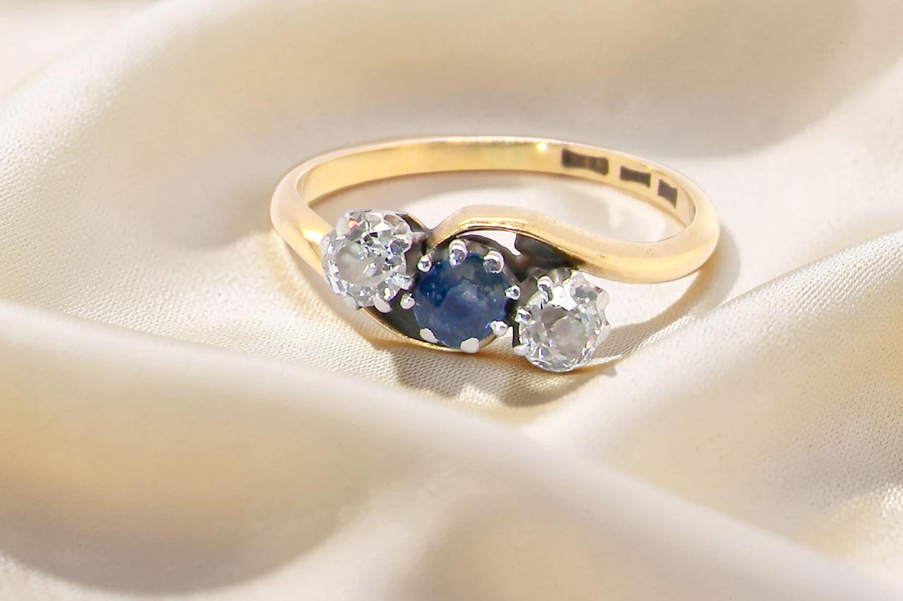 An 18 carat gold three stone cross over ring.
Centre stone a round sapphire 4mm diameter. Each side a brilliant cut white diamond 4mm diameter (0.25 carats estimated),