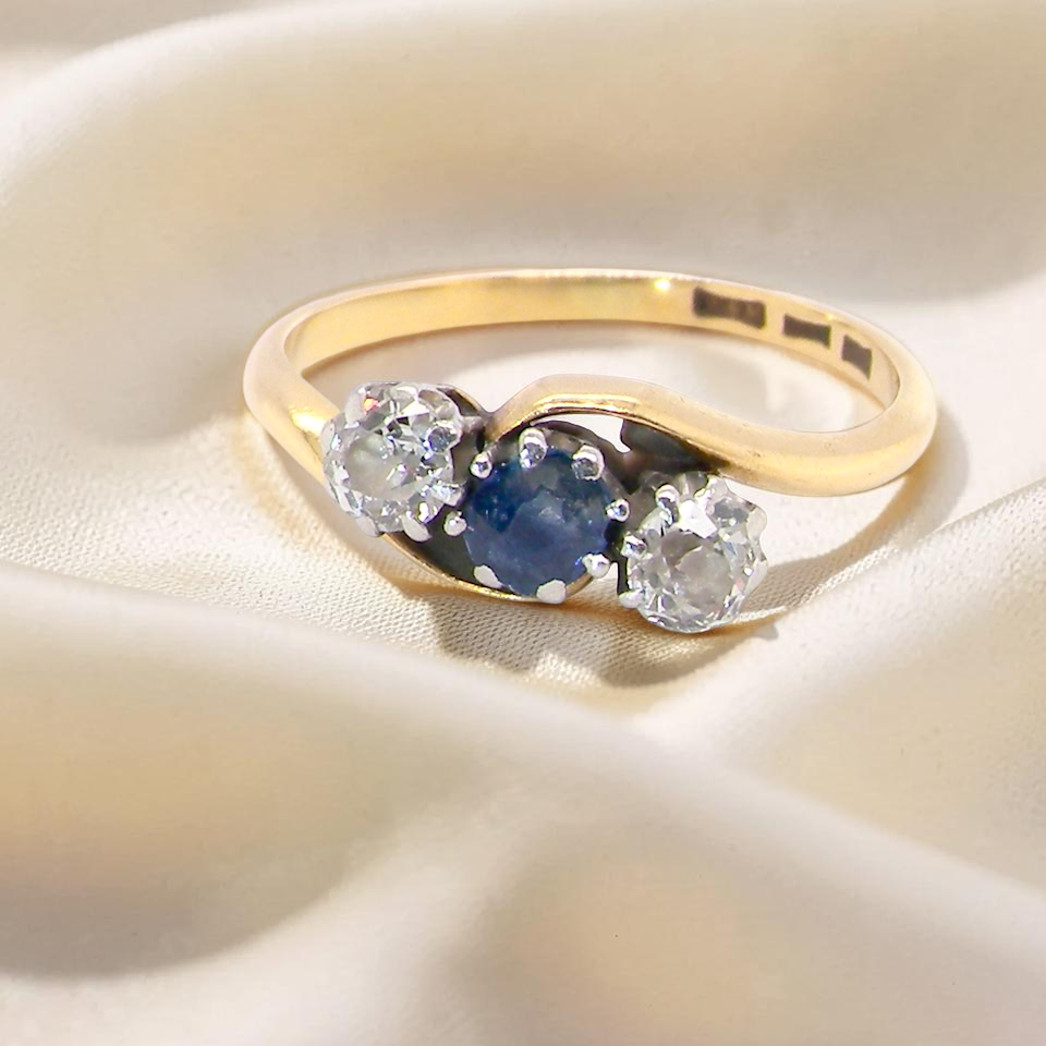 An 18 carat gold three stone cross over ring.
Centre stone a round sapphire 4mm diameter. Each side a brilliant cut white diamond 4mm diameter (0.25 carats estimated),