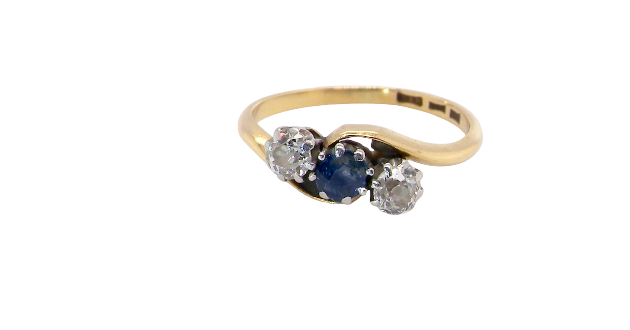 An 18 carat gold three stone cross over ring.
Centre stone a round sapphire 4mm diameter. Each side a brilliant cut white diamond 4mm diameter (0.25 carats estimated), colour i-k, clarity Si.