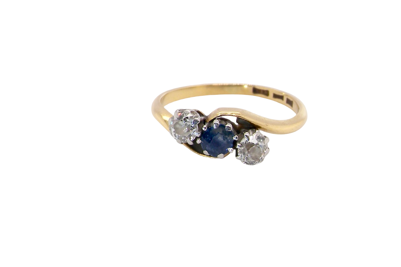 An 18 carat gold three stone cross over ring.
Centre stone a round sapphire 4mm diameter. Each side a brilliant cut white diamond 4mm diameter (0.25 carats estimated), colour i-k, clarity Si.