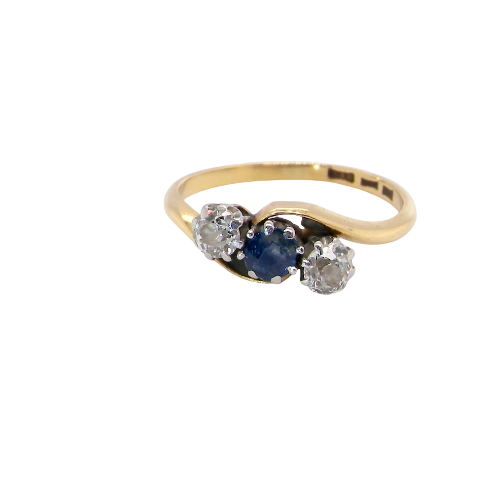 An 18 carat gold three stone cross over ring.
Centre stone a round sapphire 4mm diameter. Each side a brilliant cut white diamond 4mm diameter (0.25 carats estimated), colour i-k, clarity Si.