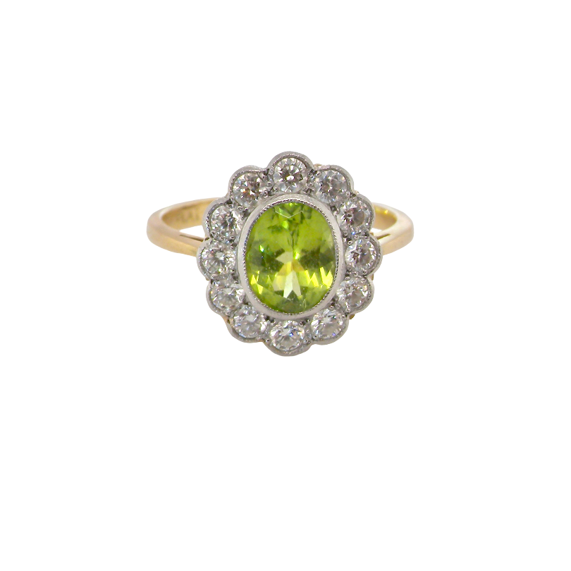 A vintage-style cluster ring featuring an oval green peridot stone surrounded by a halo of small clear stones, set on a yellow gold band, isolated on a white background.