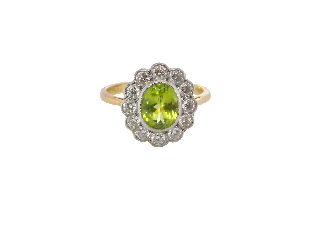 A vintage-style cluster ring featuring an oval green peridot stone surrounded by a halo of small clear stones, set on a yellow gold band, isolated on a white background.