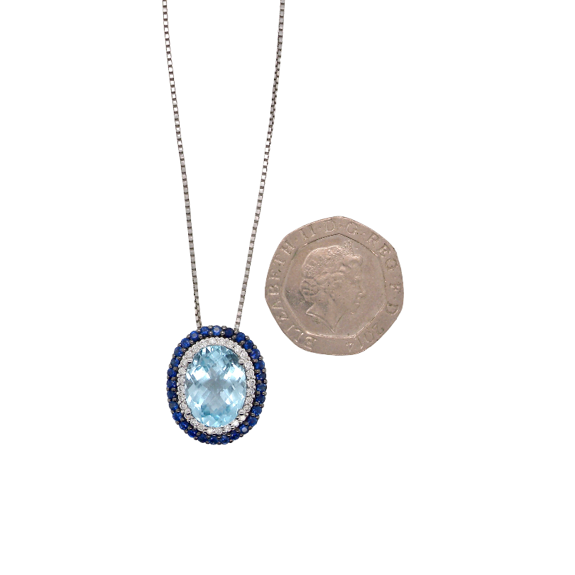 An oval pendant necklace with a light blue central stone and a halo of small dark blue stones, shown on a silver-toned chain next to a UK ten-pound coin for scale, on a plain white background.