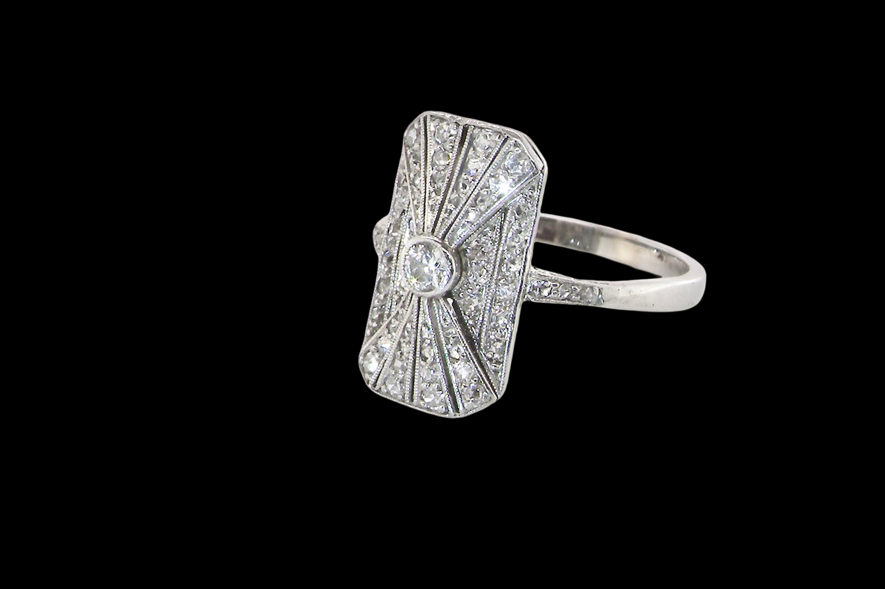 An Art Deco panel shaped cluster ring. The head measures 16x10mm, depth 3.4mm, and has two fan shaped designs that feature above and below a brilliant cut white diamond centre stone which measures (0.15 carats est)