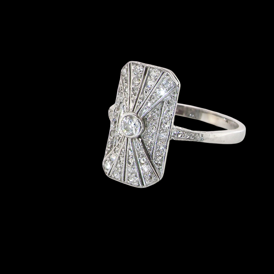 An Art Deco panel shaped cluster ring. The head measures 16x10mm, depth 3.4mm, and has two fan shaped designs that feature above and below a brilliant cut white diamond centre stone which measures (0.15 carats est)