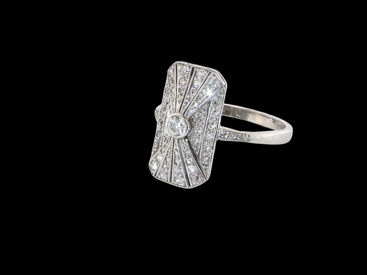 An Art Deco panel shaped cluster ring. The head measures 16x10mm, depth 3.4mm, and has two fan shaped designs that feature above and below a brilliant cut white diamond centre stone which measures (0.15 carats est)