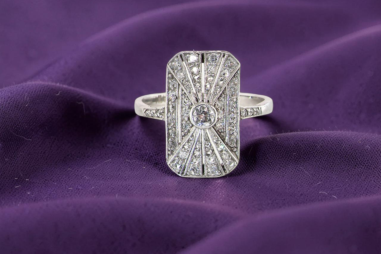 An Art Deco panel shaped cluster ring. The head measures 16x10mm, depth 3.4mm, and has two fan shaped designs that feature above and below a brilliant cut white diamond centre stone which measures 3.4mm