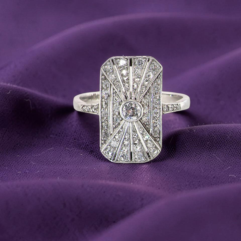 An Art Deco panel shaped cluster ring. The head measures 16x10mm, depth 3.4mm, and has two fan shaped designs that feature above and below a brilliant cut white diamond centre stone which measures 3.4mm