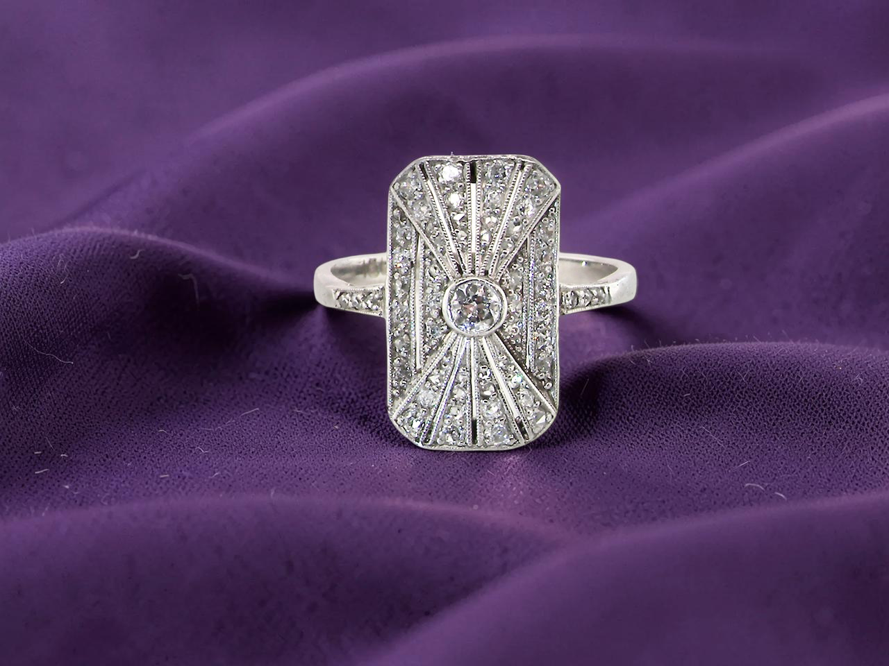 An Art Deco panel shaped cluster ring. The head measures 16x10mm, depth 3.4mm, and has two fan shaped designs that feature above and below a brilliant cut white diamond centre stone which measures 3.4mm