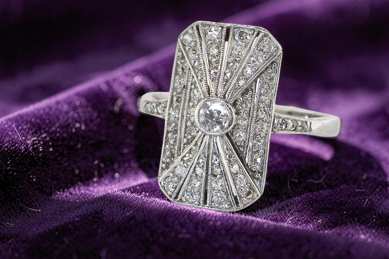 An Art Deco panel shaped cluster ring. The head measures 16x10mm, depth 3.4mm, and has two fan shaped designs that feature above and below a brilliant cut white diamond centre which measures 3.4mm (0.15 carats est)