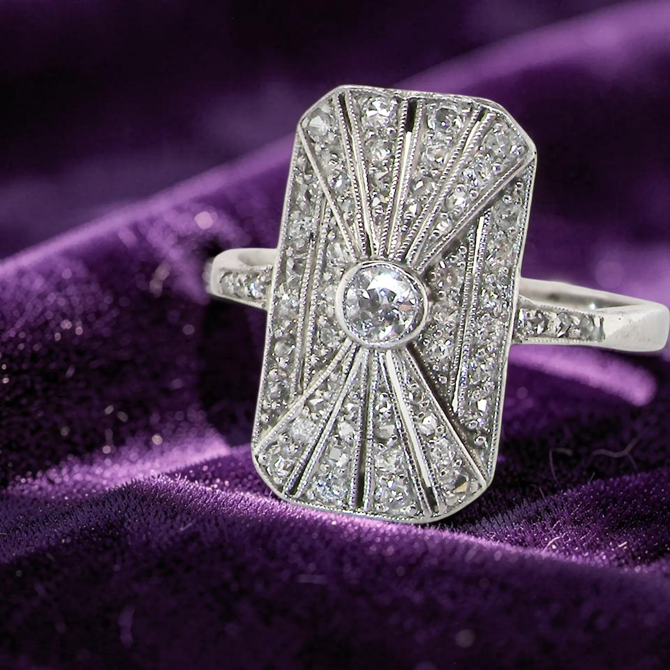 An Art Deco panel shaped cluster ring. The head measures 16x10mm, depth 3.4mm, and has two fan shaped designs that feature above and below a brilliant cut white diamond centre which measures 3.4mm (0.15 carats est)