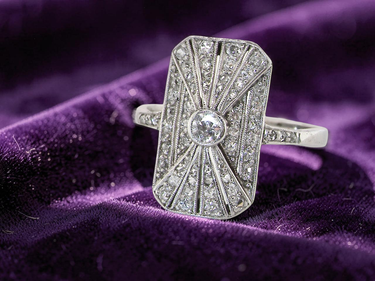 An Art Deco panel shaped cluster ring. The head measures 16x10mm, depth 3.4mm, and has two fan shaped designs that feature above and below a brilliant cut white diamond centre which measures 3.4mm (0.15 carats est)