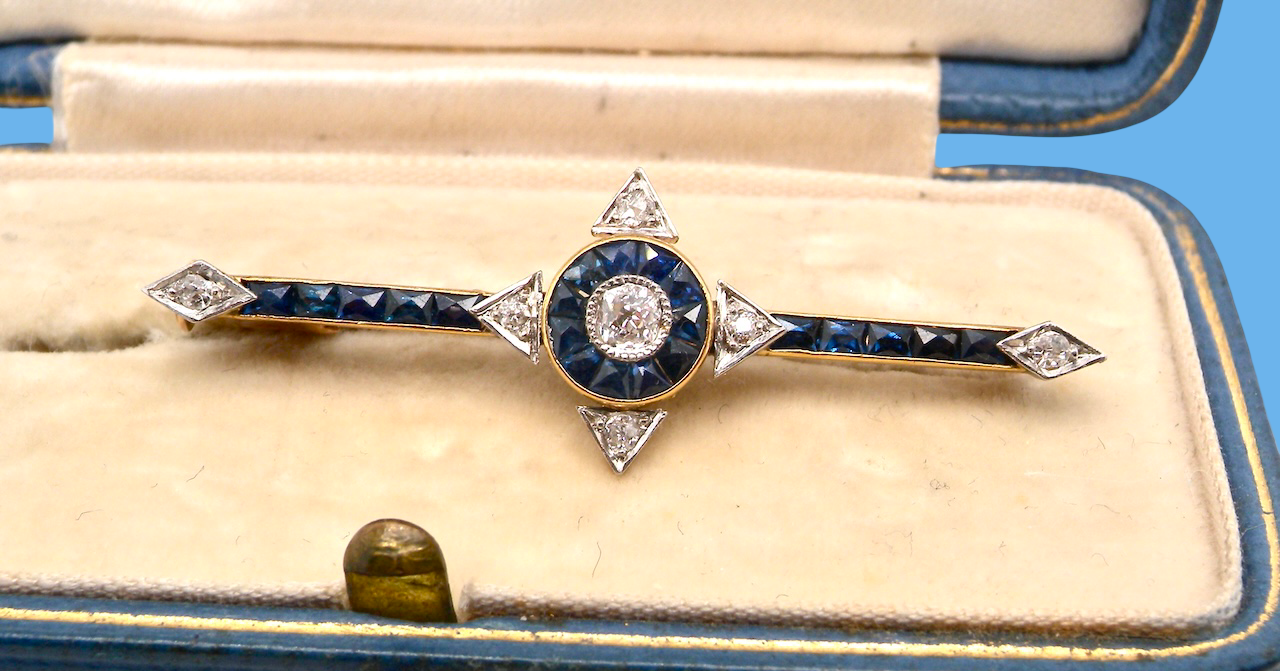 An Edwardian bar brooch. The centre 'target' style portion is set with a brilliant cut white diamond 4mm (0.25 carats) colour H-j, . Mille grain setting.
