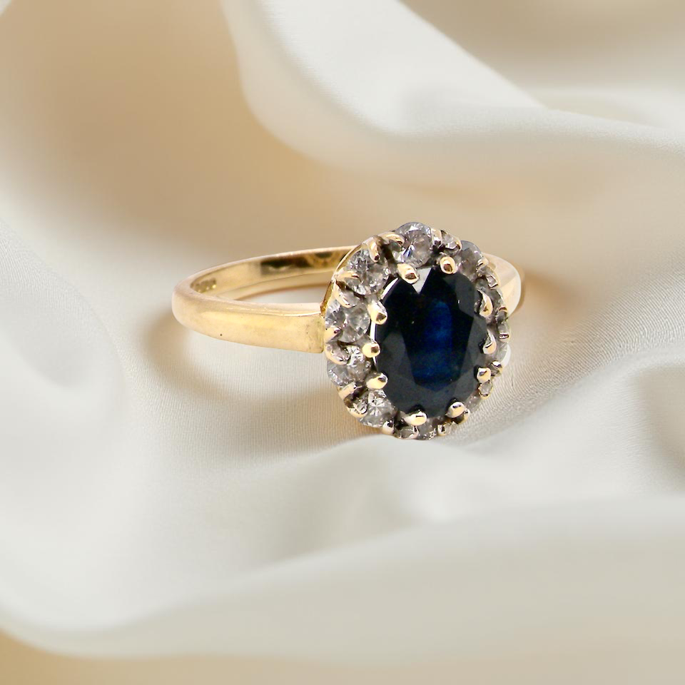 An elegant 18 carat gold cluster ring featuring a striking inky dark blue sapphire at its centre. B