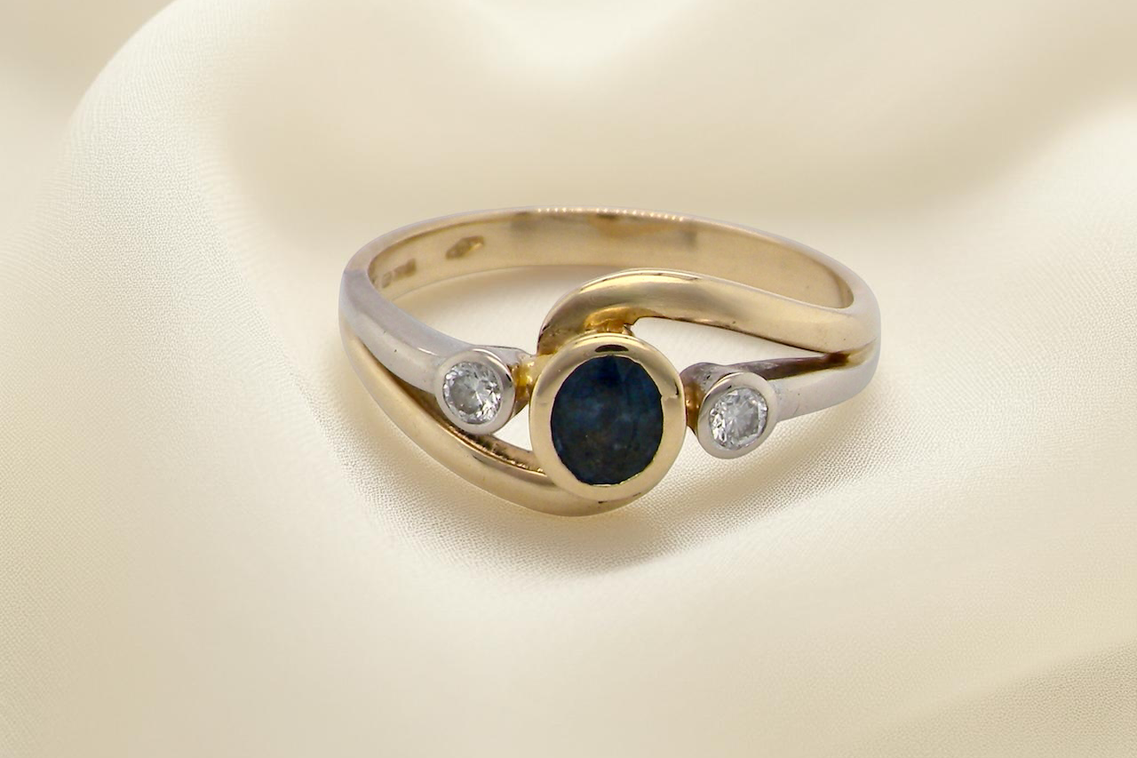 An elegant 18 carat gold three-stone ring featuring a rich, oval-cut dark blue sapphire at the centre, measuring 5 × 4mm, secured in a classic rim setting.