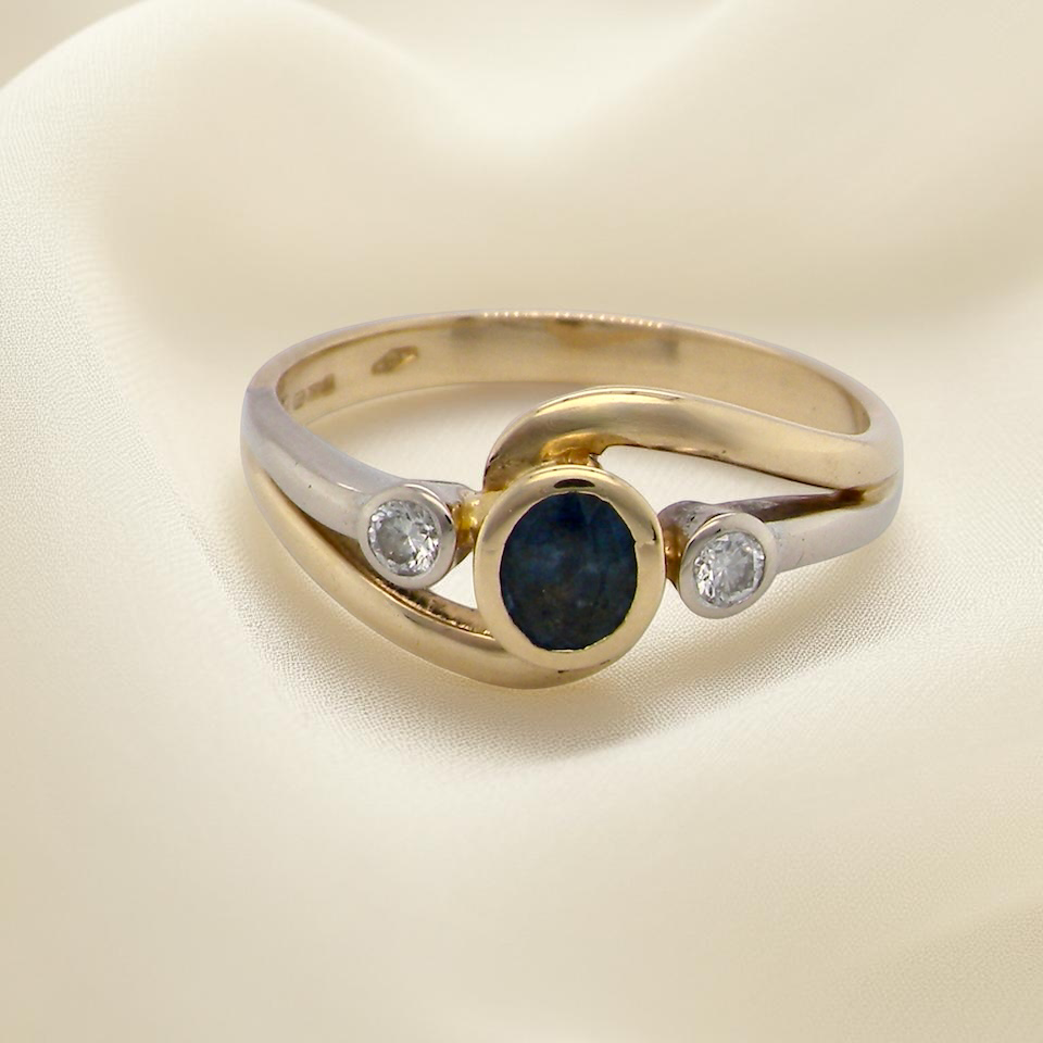 An elegant 18 carat gold three-stone ring featuring a rich, oval-cut dark blue sapphire at the centre, measuring 5 × 4mm, secured in a classic rim setting.
