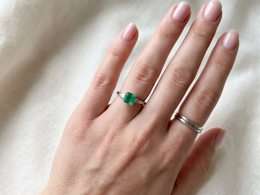 An elegant platinum ring featuring a stunning step-cut emerald to the centre, measuring approximately 5.5 x 5.5mm and estimated at 0.80 carats. The emerald displays a vibrant, deep green colour with excellent presence.