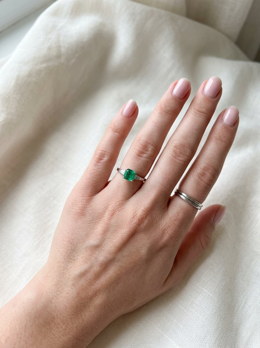 An elegant platinum ring featuring a stunning step-cut emerald to the centre, measuring approximately 5.5 x 5.5mm and estimated at 0.80 carats. The emerald displays a vibrant, deep green colour with excellent presence.
