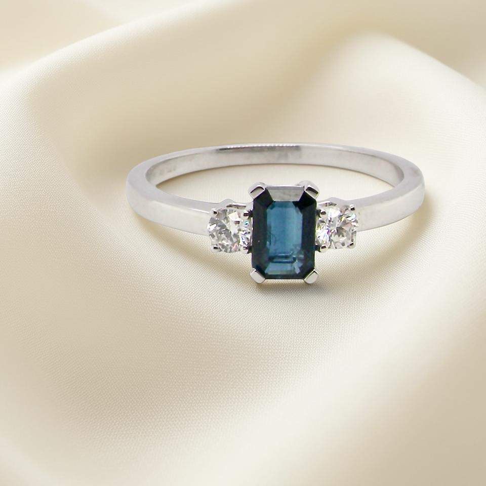 An elegant three-stone ring crafted in 18 carat white gold, featuring a striking rectangular dark blue sapphire at the centre, measuring 6 × 4 mm